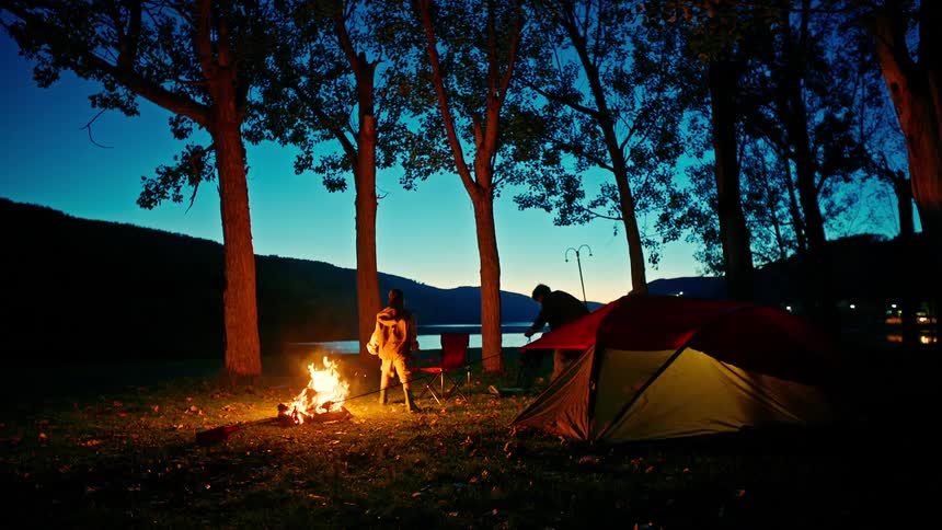 Two brothers move around a burning fire at a campsite before sitting down in chairs to take in the view