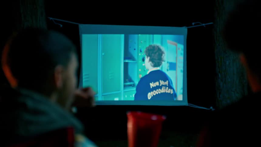 Two people watch a movie projected on a sheet at night while one eats from a red cup