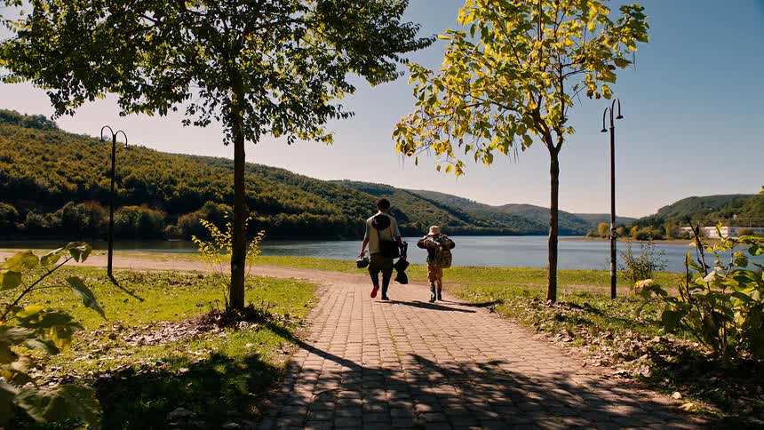 A man and young boy walk down a paved path toward a lake carrying fishing gear