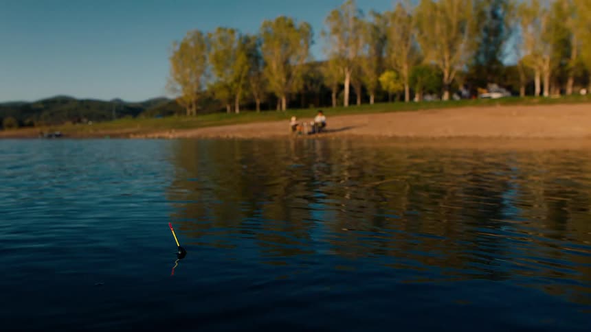 A fishing bob disappears under a lake where two people are fishing in the shore