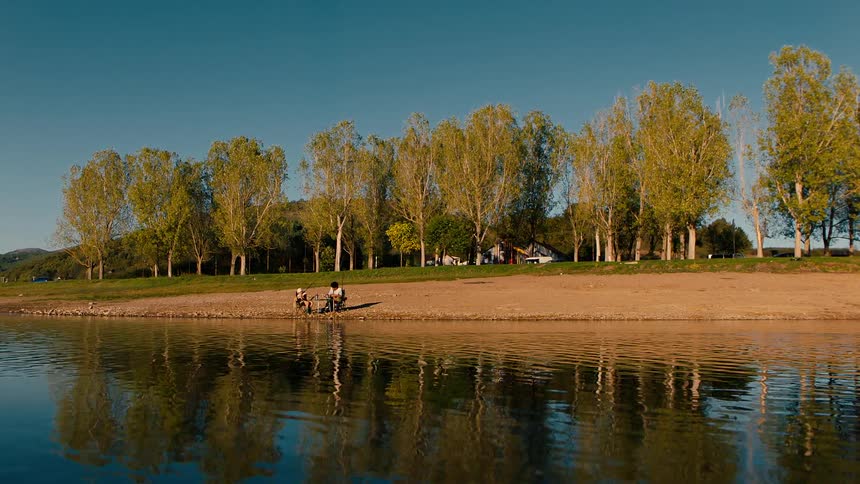 Two people sit on a dirt area on the shore of a lake