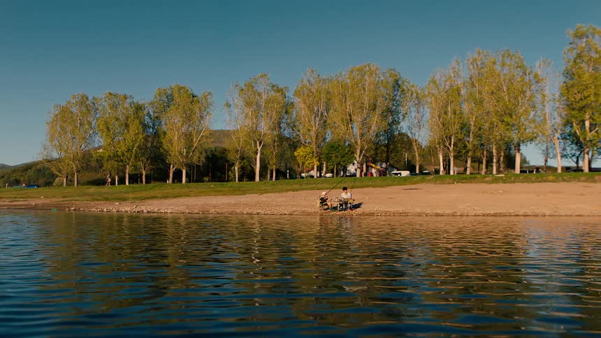 Two people sit on a lakeshore and fish