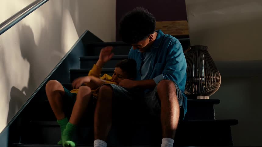 A young boy sits sadly on the stairs until an older boy comes comforts him and they walk down the stairs together