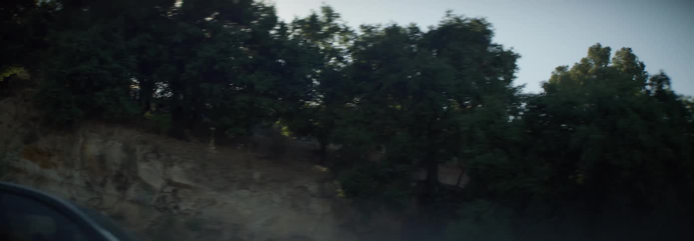 A person drives a small car on a road next to a hillside on a sunny summer day