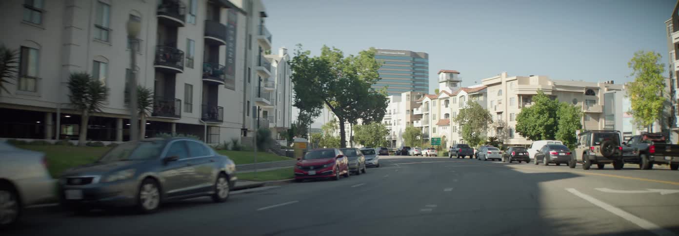 A bustling city street lined with apartment buildings and parked cars with a glimpse of a taller skyscraper in the distance