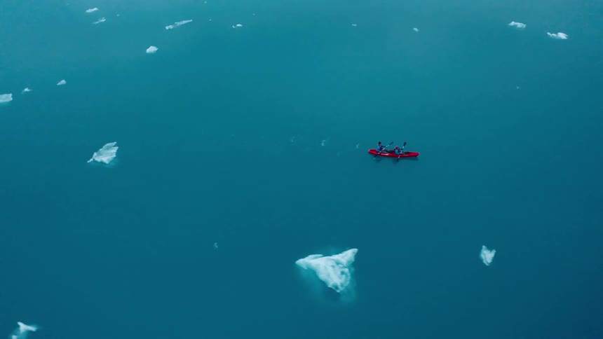 Two people paddle a red kayak through blue water dotted with small icebergs