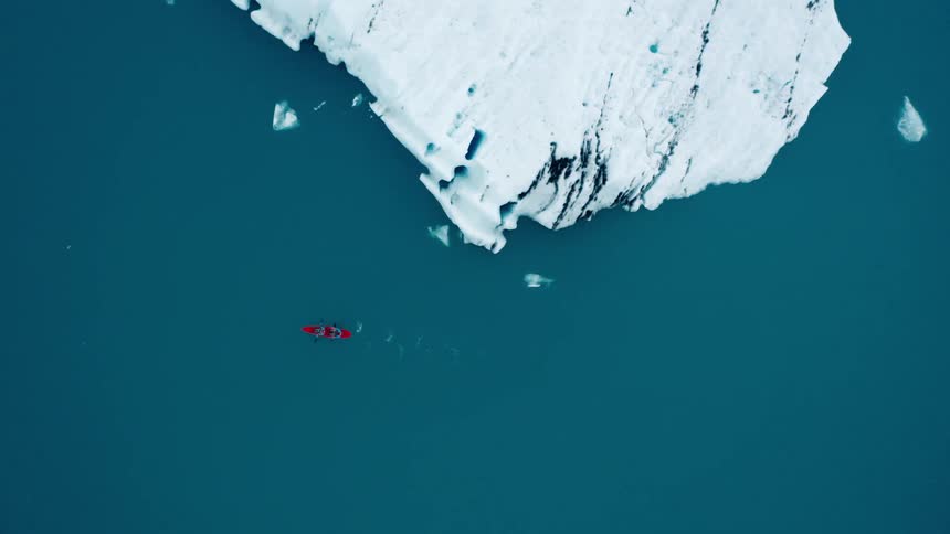 A boat with two people in it rows past a large iceberg