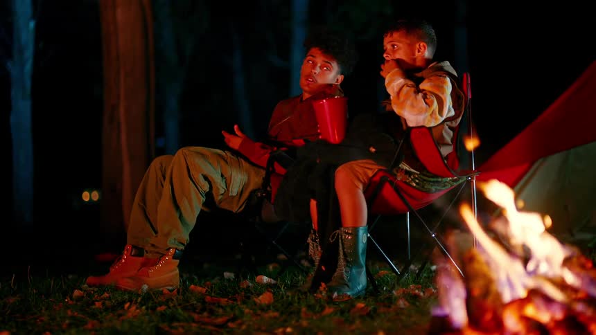Siblings eat and talk while sitting near a campfire at night