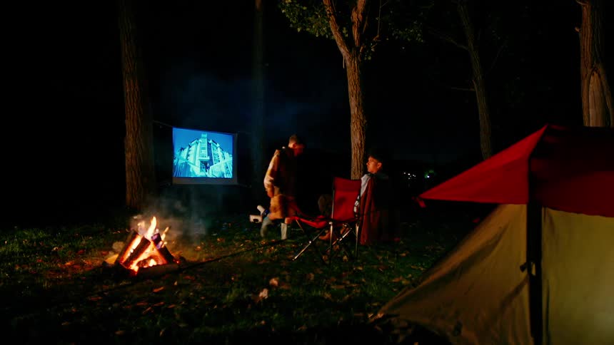 Younger brother adjusts a projector while he and his older sibling watch movies outdoors at their campsite at night