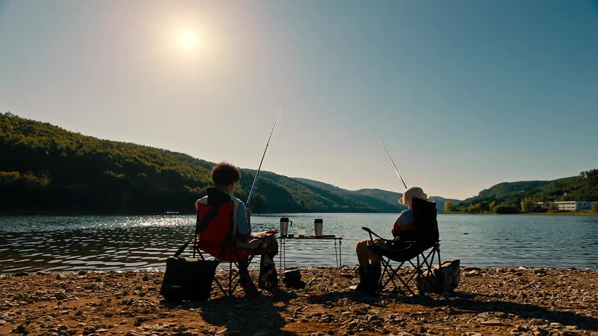Siblings sit in chairs on the lake shore and fish on a sunny summer day