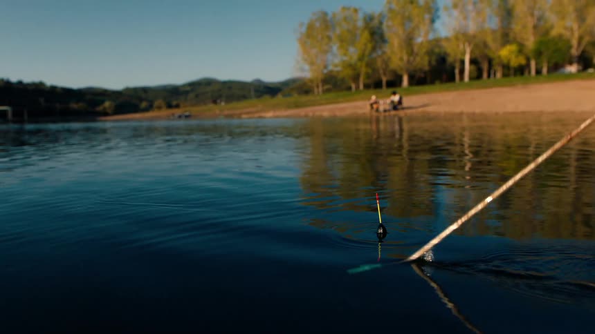 A person pulls a fishing line out of a lake as two people sit nearby on shore