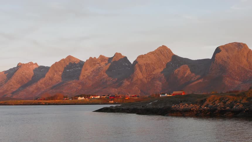The sun casts a golden glow on a coastal town nestled amidst a dramatic mountain range