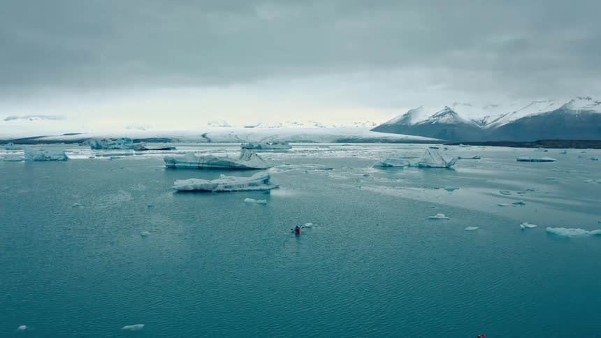 Vast body of water with floating icebergs and a small red boat in the center with distant mountains in the background under a cloudy sky