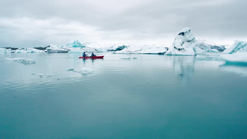 Two people kayak through icy blue waters dotted with icebergs and glaciers under a cloudy sky
