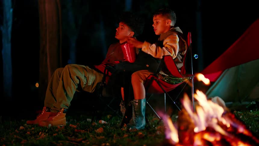 Two young men sitting in camping chairs next to a campfire at night