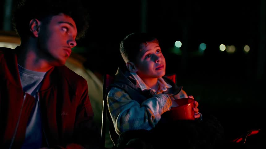 Two young boys sitting outdoors at night illuminated by a outdoor tv one is holding a mug and both are looking up with expressions of wonder or fascination