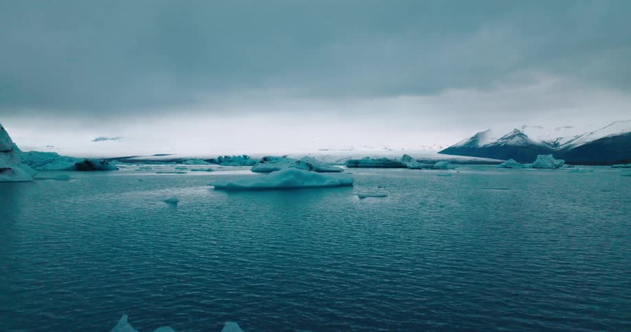 A vast expanse of icy blue water stretches towards the horizon with floating icebergs and framed by snow capped mountains