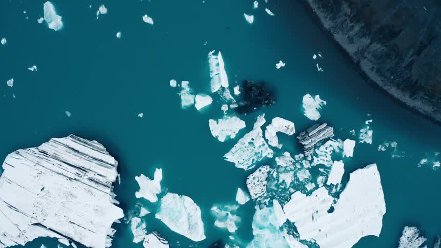 Icebergs and smaller pieces of ice float in the water off a shore during the day