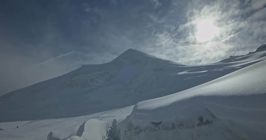 A snowy mountainous landscape stretches into the distance partially obscured by clouds and lit by the sun