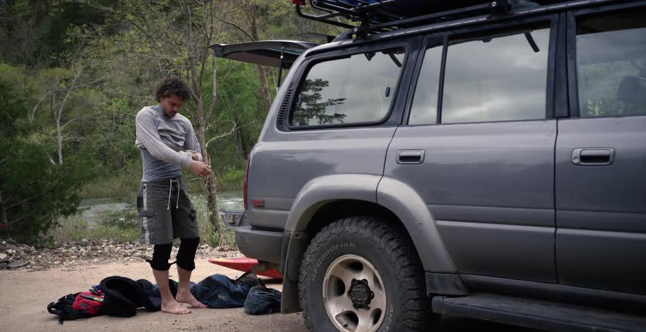 A man starts to remove his shirt behind his car