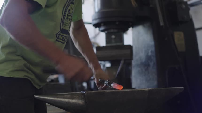A blacksmith carefully shapes a glowing piece of metal on an anvil using a pair of tongs and a hammer