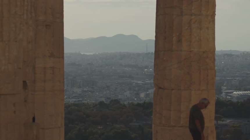 A man walking between two large ancient stone columns with a sprawling cityscape in the background framed by distant hills under a hazy sky
