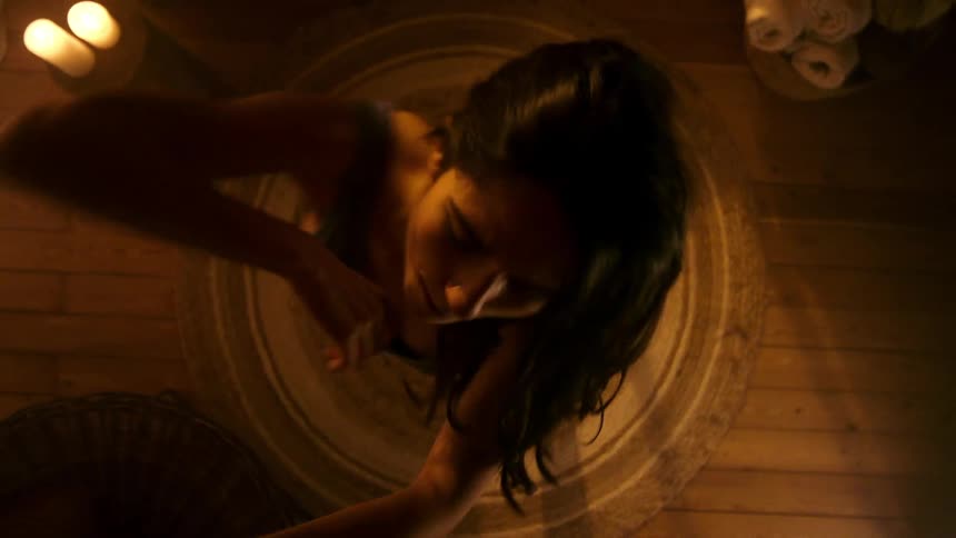 A woman dancing joyfully in a dimly lit cozy room with wooden floors surrounded by warm candlelight and rolled towels as seen from an overhead angle