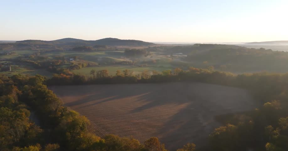 The sun casts a golden glow over the rolling hills where fields of brown stubble meet patches of vibrant autumn foliage