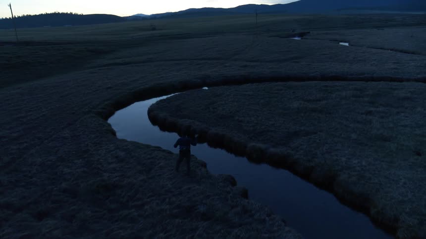 A lone figure stands on a riverbank at dusk overlooking a winding river