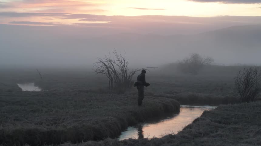 A lone fisherman casts a line into a misty river at dawn