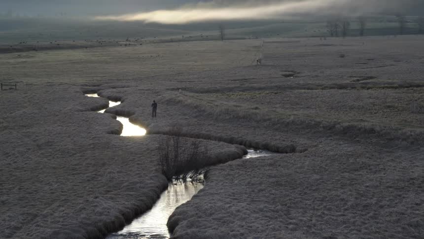 A lone figure walks along a winding river through a misty barren landscape