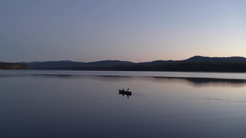 Rowboat traveling across a calm lake surrounded by mountains on a morning with a purple sky