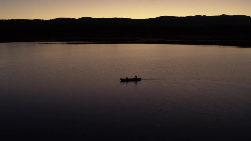 Silhouette of two people in a canoe on a lake during sunrise with the horizon marked by hills or low mountains