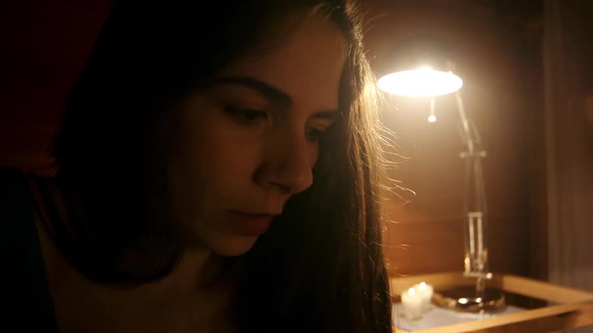 Close up of the face of a woman with long hair sitting on a room next to a table lamp