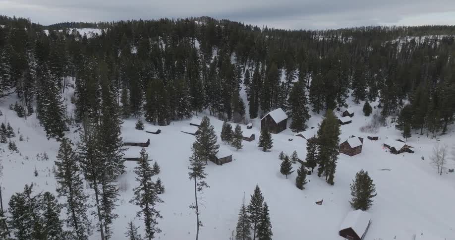 A blanket of snow covers the rustic cabins and the surrounding forest creating a serene winter scene