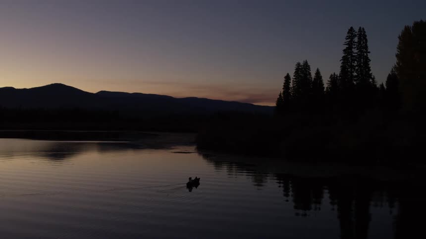 Serene scene of two people in a small boat on a calm body of water with a forest and mountains silhouetted against a twilight sky