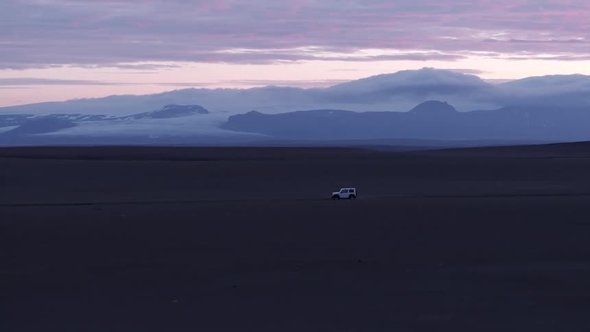 A white jeep travels along a remote road near a mountain range at sunset