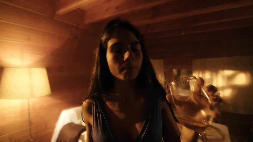 A young woman holding a glass of wine walks drunkenly in a cozy bedroom