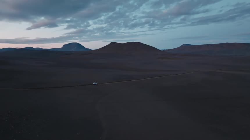 Aerial view of a white car traveling along a road surrounded by black soil with mountains on the horizon