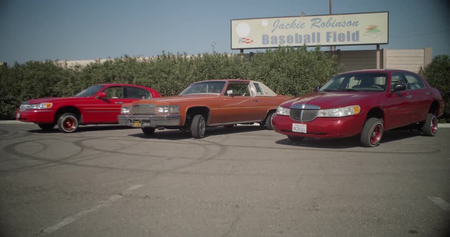 Three lowrider cars parked in a parking lot in front of a baseball field