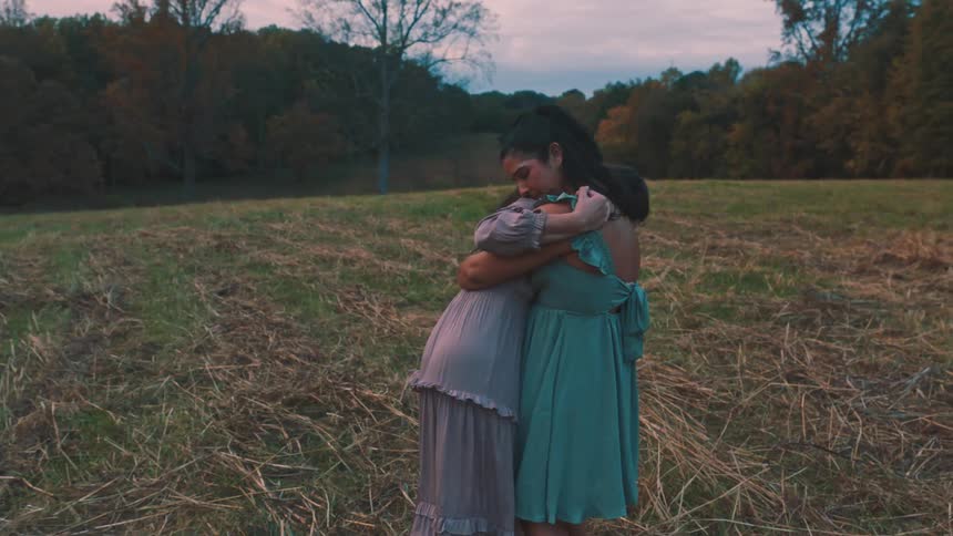 Two women in dresses hugging each other in a field in a melancholic dance