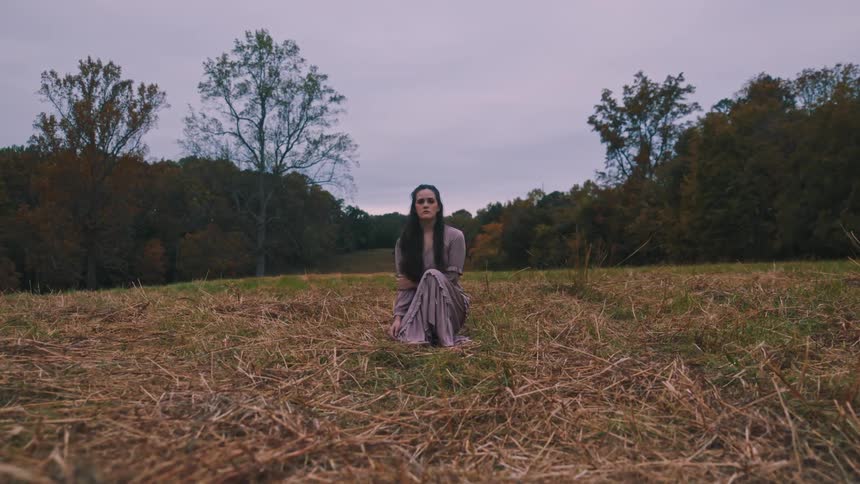 Woman in a light purple dress crouching in a field surrounded by trees