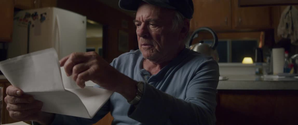 Elderly man wearing a cap and longsleeved shirt is sitting at the kitchen table examining a document with a serious expression