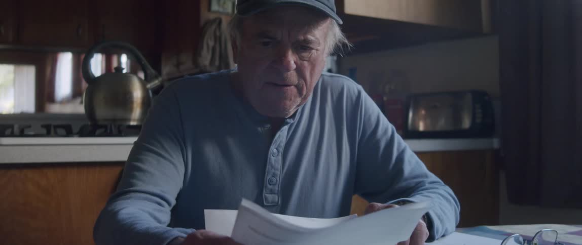 An elderly man sits down in the kitchen smiling as he reads a letter