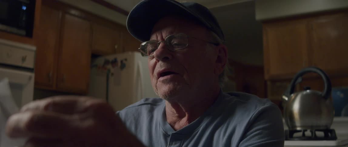 An elderly man sits in his kitchen laughing as he opens a letter