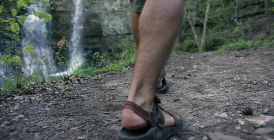Man walking a dirt path alongside a waterfall as seen from his knees down