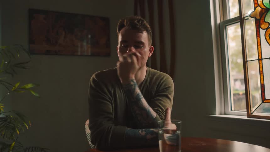 A tattooed young man sits on a small table pondering in front of a glass of water