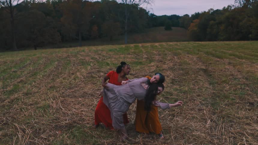 Three women in colorful dresses dancing in a performance in a pasture surrounded by trees