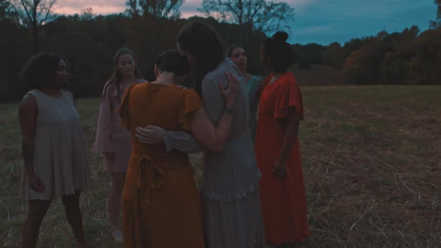 Group of women in different colored dresses holding hands and hugging each other and squatting in a field in a performance