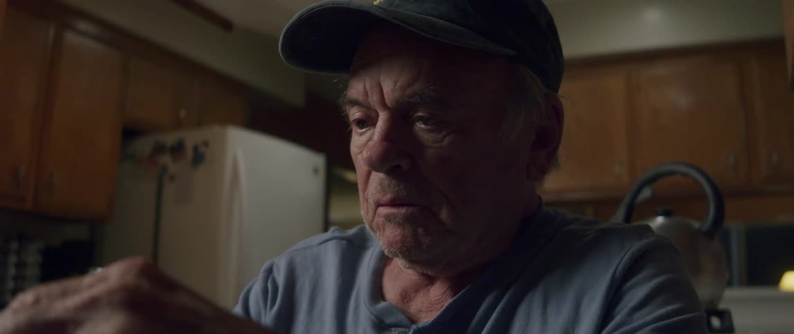 An elderly man sits in his kitchen laughing as he opens a letter